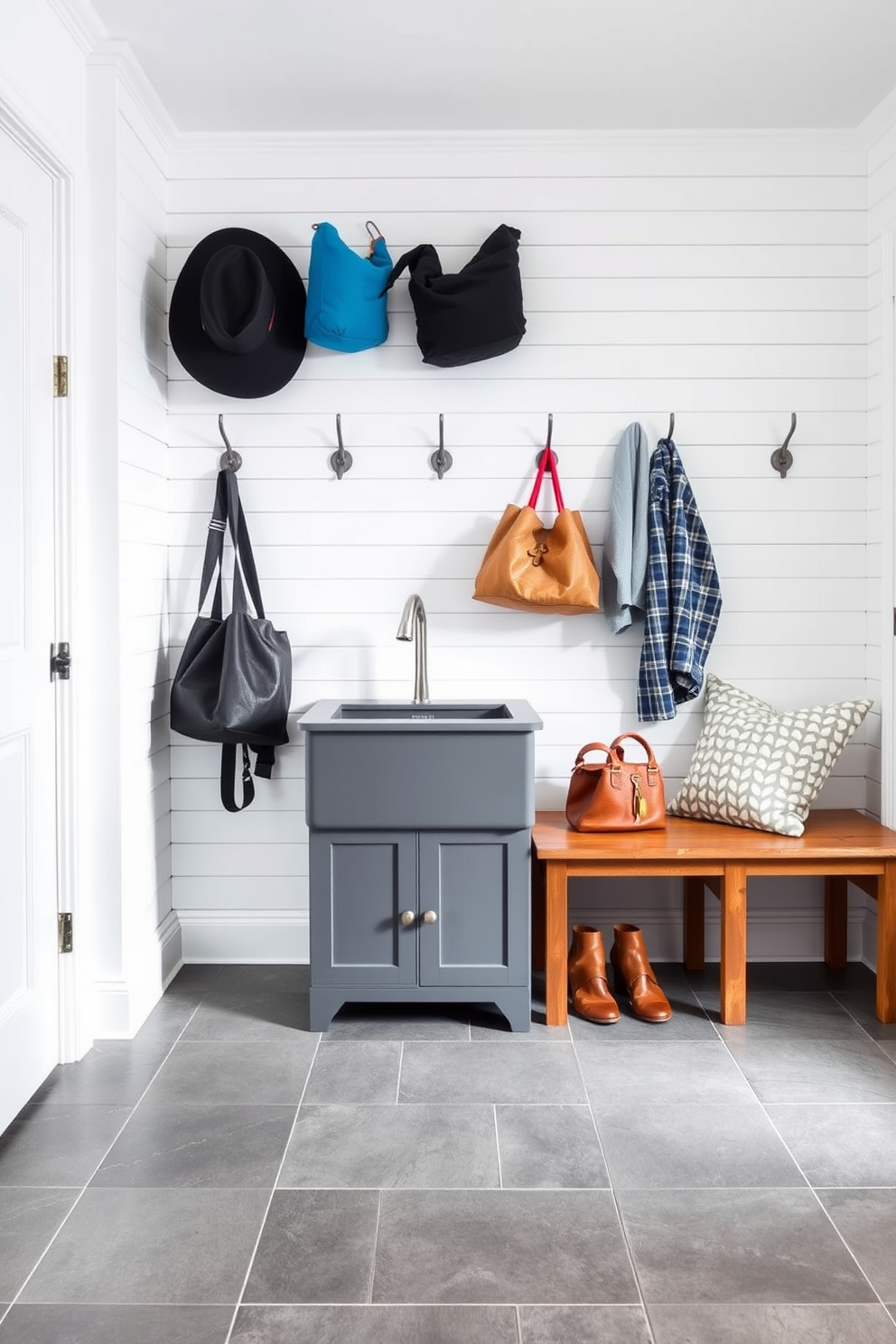 A stylish mudroom featuring a gray utility sink set against a backdrop of shiplap walls painted in a soft white. The floor is adorned with large gray tiles, and a row of hooks above a wooden bench holds coats and bags, creating an organized and inviting space.