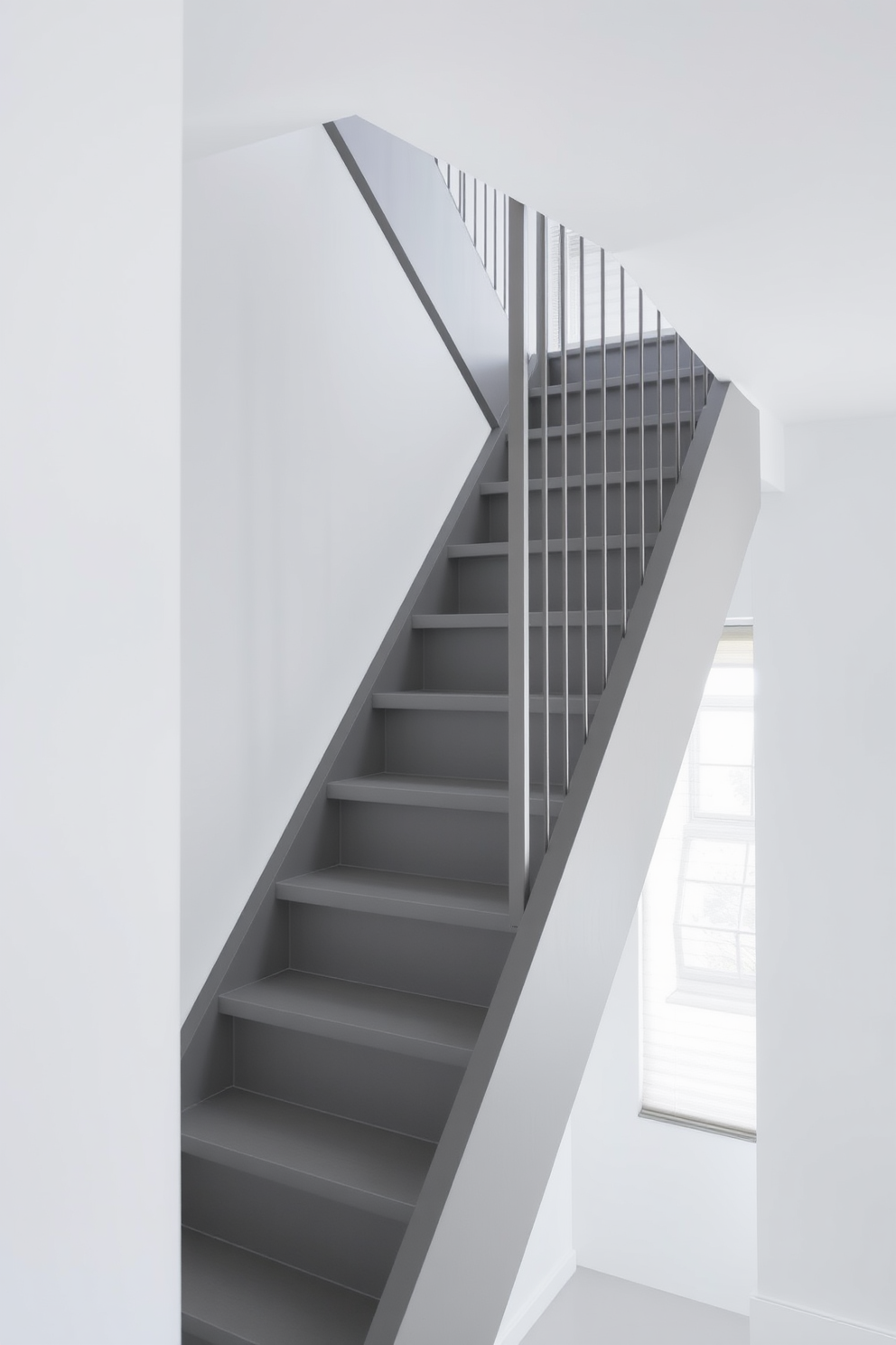 A dark gray staircase with a minimalist design showcases clean lines and a sleek appearance. The steps are made of polished concrete, complemented by a simple metal railing that enhances the modern aesthetic. Natural light filters in through a large window, casting soft shadows on the staircase. The surrounding walls are painted in a crisp white, creating a striking contrast with the dark gray tones.