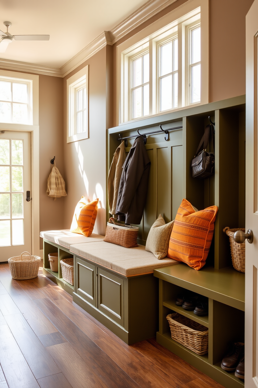 A spacious mudroom features olive green built-in storage benches that provide ample seating and organization. The benches are adorned with plush cushions in complementary colors, creating a cozy and inviting atmosphere. Natural light floods the space through large windows, highlighting the earthy tones of the walls and floor. The mudroom is designed with practical elements such as hooks for coats and baskets for shoes, ensuring functionality meets style.