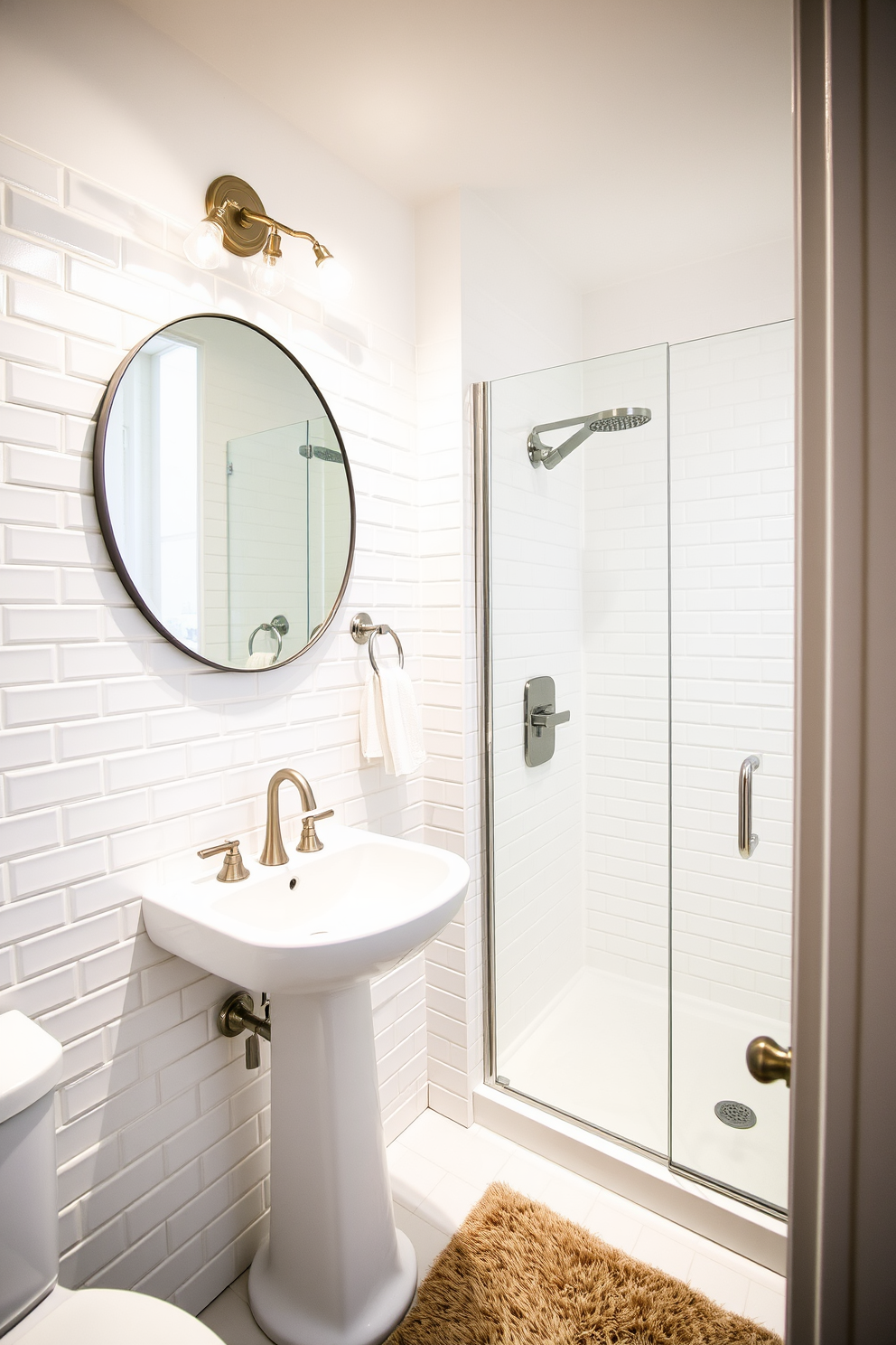 A stylish guest bathroom featuring beveled white subway tile walls that create a clean and timeless look. The space includes a sleek pedestal sink with a brushed nickel faucet and a large round mirror that enhances the room's brightness. A comfortable shower area with a glass enclosure showcases a rainfall showerhead and minimalist shelving for toiletries. Soft lighting fixtures above the sink and near the shower add warmth, while a plush bath mat complements the overall aesthetic.