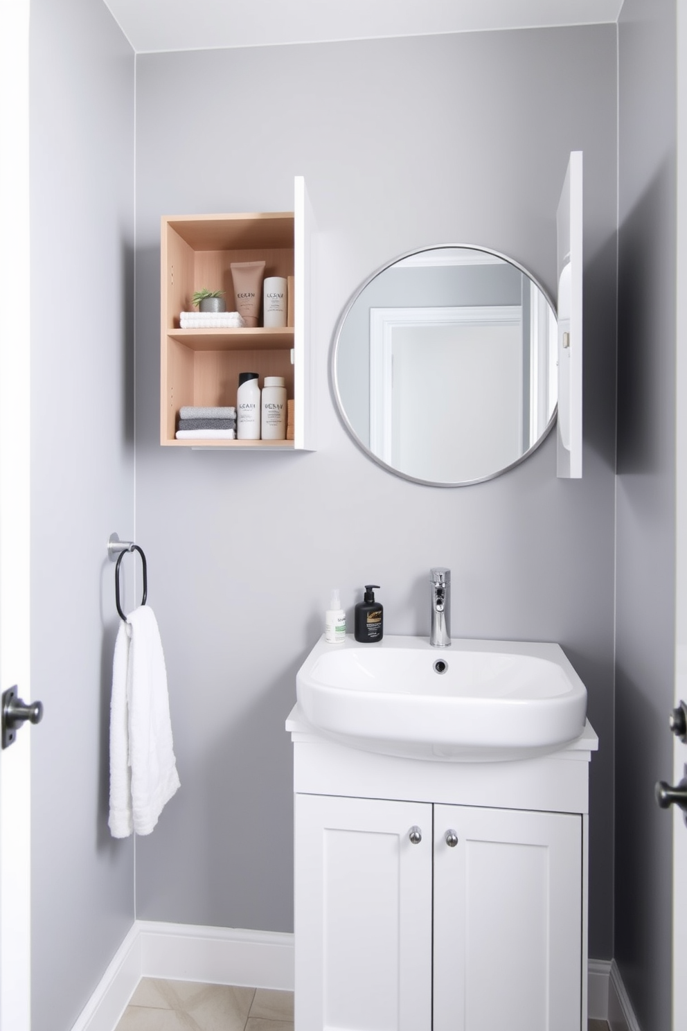 A stylish half bathroom featuring compact storage solutions that maximize space. A sleek wall-mounted cabinet with open shelving holds neatly arranged toiletries and decorative items. The color palette consists of soft gray walls paired with a white vanity that includes hidden drawers. A round mirror with a minimalist frame hangs above the sink, enhancing the room's modern aesthetic.