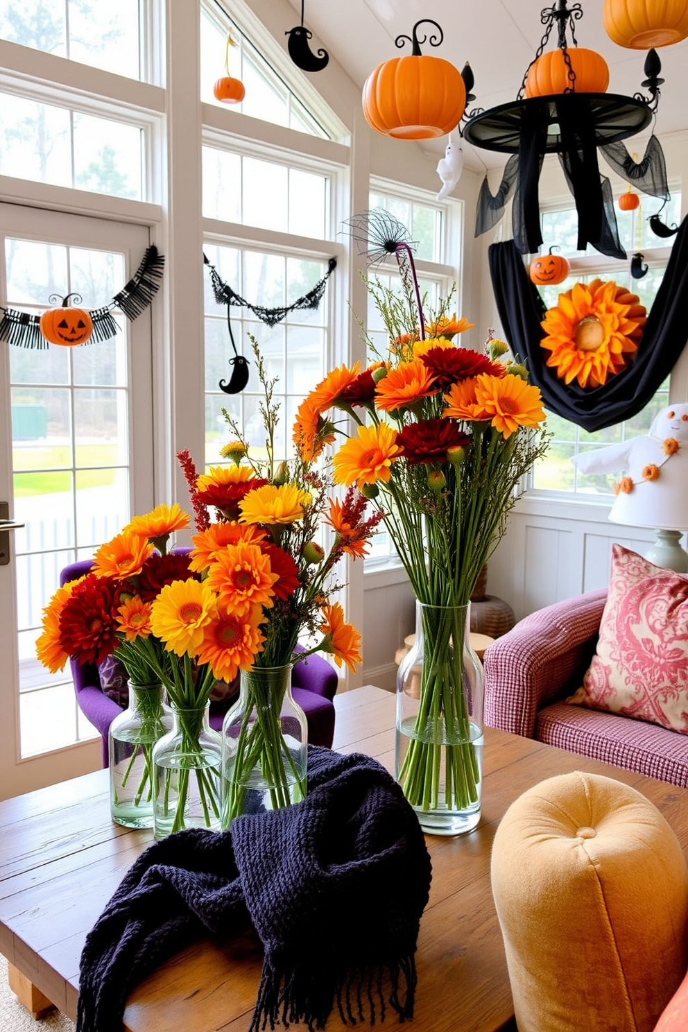 Colorful fall flowers in vases are arranged on a rustic wooden table in a sunroom filled with natural light. The vibrant blooms in shades of orange, yellow, and red contrast beautifully with the warm, earthy tones of the sunroom's decor. Halloween decorations adorn the sunroom, with playful touches like miniature pumpkins and whimsical ghosts hanging from the ceiling. Soft, cozy throws in deep purple and black are draped over the furniture, creating a festive yet inviting atmosphere.