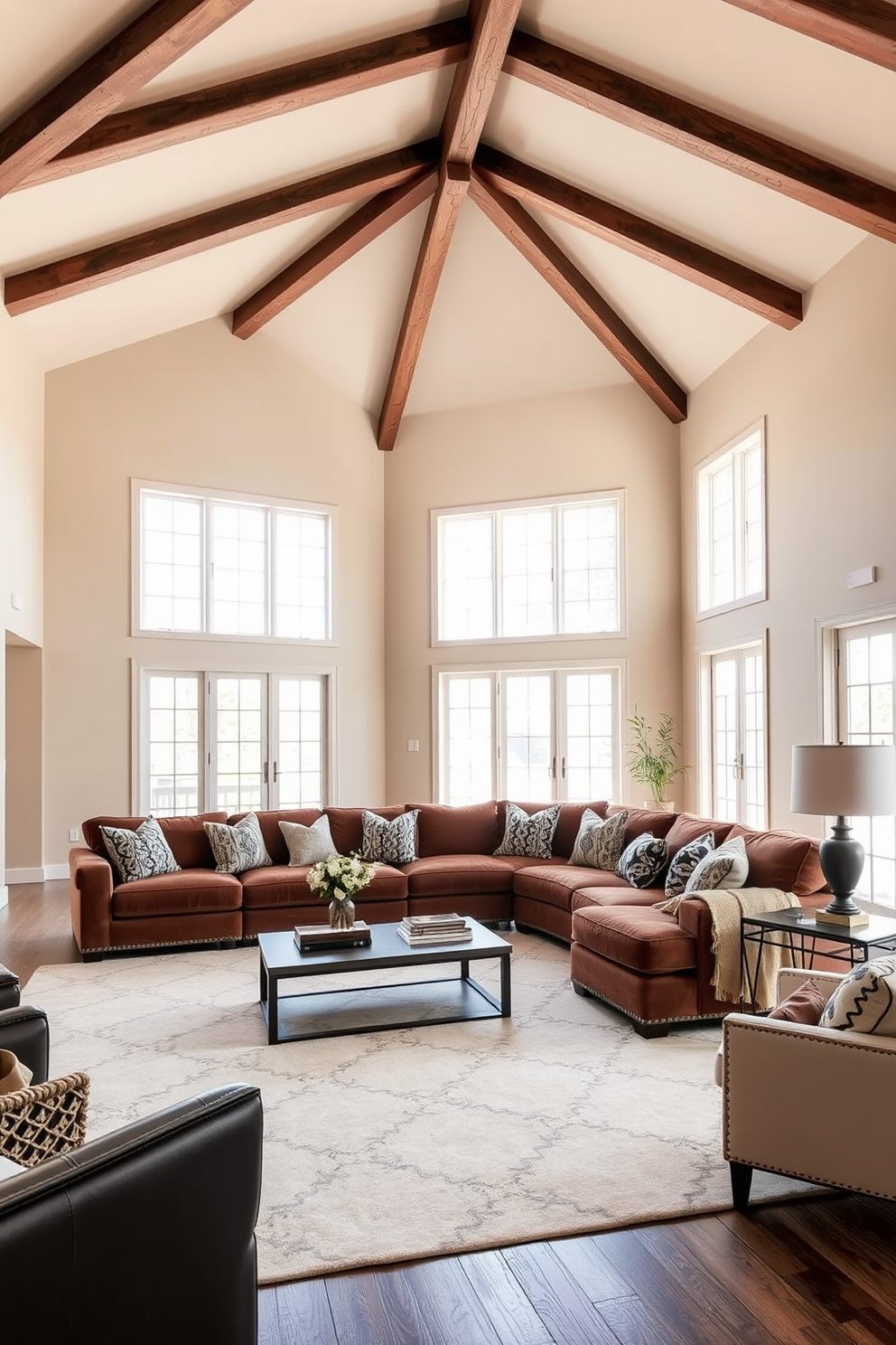 A spacious living room with a high ceiling featuring exposed wooden beams. The walls are painted in a soft beige tone, creating a warm backdrop for a large sectional sofa upholstered in a rich velvet fabric. A large area rug with a subtle geometric pattern anchors the seating area, adding depth and texture. Floor-to-ceiling windows allow natural light to flood the space, highlighting a mix of decorative pillows and throws in varying fabrics and colors.