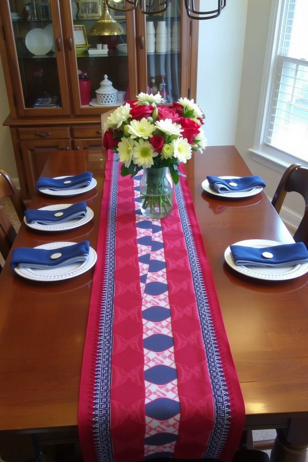 A patriotic dining room setting features a table adorned with a vibrant table runner showcasing stars and stripes in red white and blue. The wooden dining table is set with matching plates and napkins while small American flags are placed in decorative holders around the table.