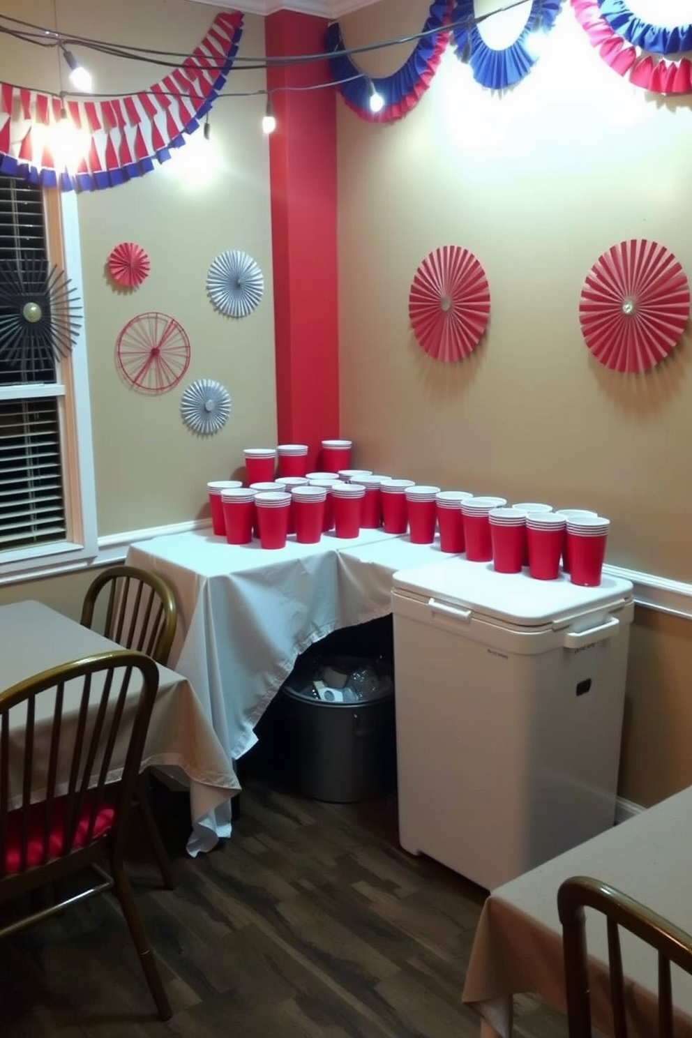 A festive drink station is set up in the corner of the dining room featuring bright red cups arranged neatly on a white tablecloth. Colorful decorations in red, white, and blue adorn the walls, creating a cheerful atmosphere for Independence Day celebrations. A large cooler filled with ice sits next to the drink station, offering a variety of refreshing beverages. String lights hang overhead, adding a warm glow to the festive setting as guests gather to enjoy the holiday.