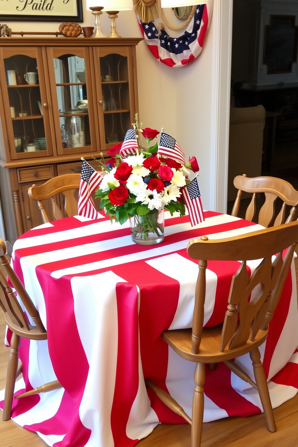 A festive dining room setting for Independence Day. The table is adorned with a red and white striped tablecloth that drapes elegantly to the floor. Surrounding the table are mismatched rustic wooden chairs, each with a unique design. Centered on the table is a vibrant bouquet of red, white, and blue flowers in a glass vase, complemented by small American flags as accents.