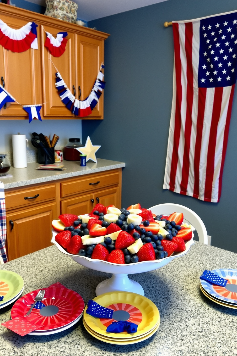 A festive kitchen setting decorated for Independence Day. The table is adorned with a vibrant red, white, and blue fruit salad, featuring strawberries, blueberries, and banana slices arranged in a star shape. Bunting in patriotic colors hangs from the cabinets, and a large American flag is displayed prominently on the wall. The countertops are topped with decorative plates and napkins that match the holiday theme, creating a cheerful and inviting atmosphere.