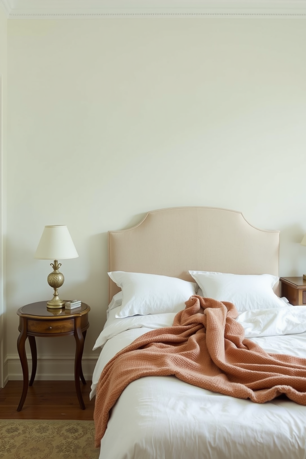 A serene Italian bedroom featuring soft muted color palettes that evoke tranquility. The walls are painted in a gentle cream shade, complemented by a plush upholstered headboard in a light taupe fabric. The bedding is layered with delicate pastel linens and a cozy throw blanket draped over the edge. Elegant wooden furniture, including a bedside table with a vintage lamp, enhances the warm and inviting atmosphere.