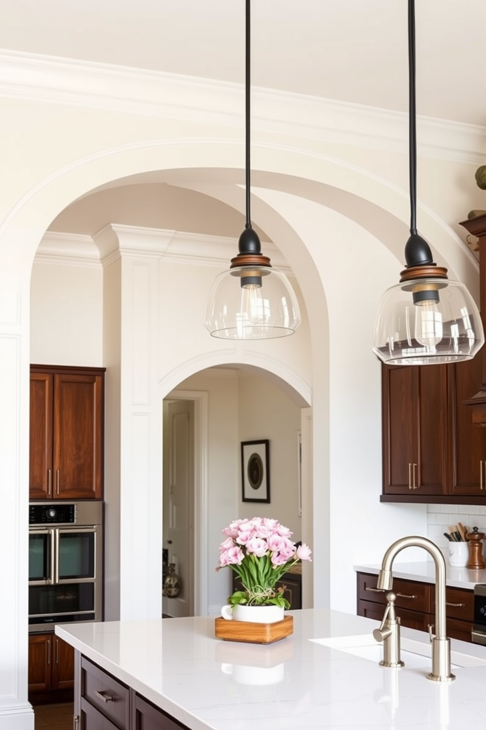 A bright and airy kitchen features an arched window above the sink, allowing natural light to flood the space. The window is framed with elegant white trim, complementing the soft pastel colors of the cabinetry. Below the window, a farmhouse-style sink is installed, surrounded by a beautiful countertop made of polished quartz. The kitchen is adorned with hanging pendant lights that add a touch of warmth and sophistication to the overall design.