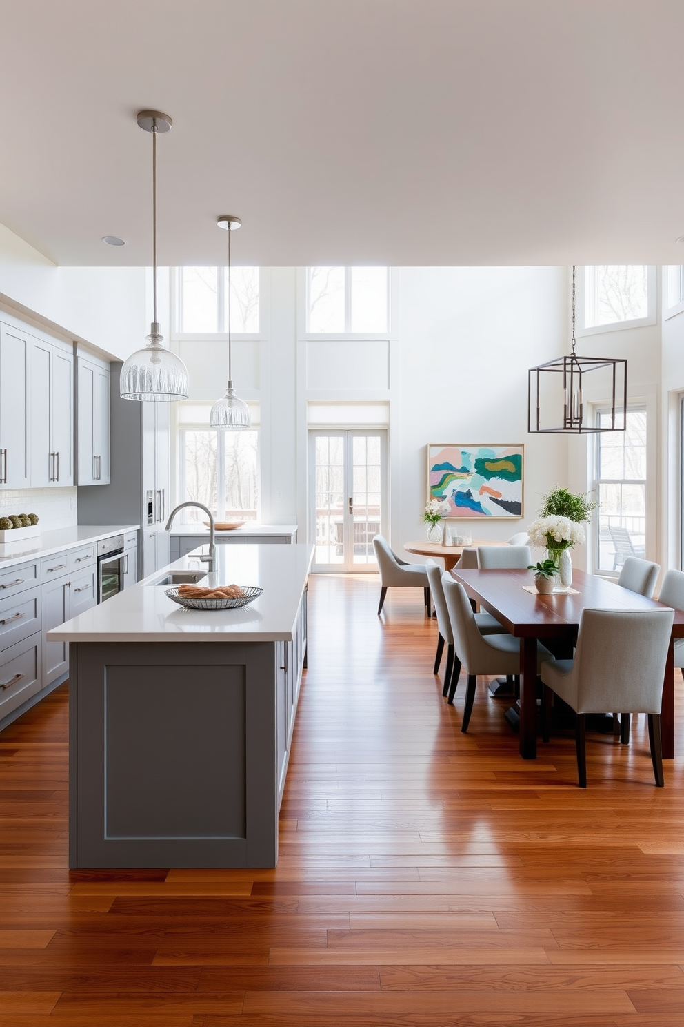 Open concept kitchen and dining room featuring a large central island with seating for four. The kitchen boasts sleek cabinetry in a soft gray finish, complemented by a white quartz countertop and modern pendant lighting above the island. The dining area includes a stylish wooden dining table surrounded by upholstered chairs, creating an inviting atmosphere. Large windows allow natural light to flood the space, highlighting the warm wood flooring and contemporary decor.