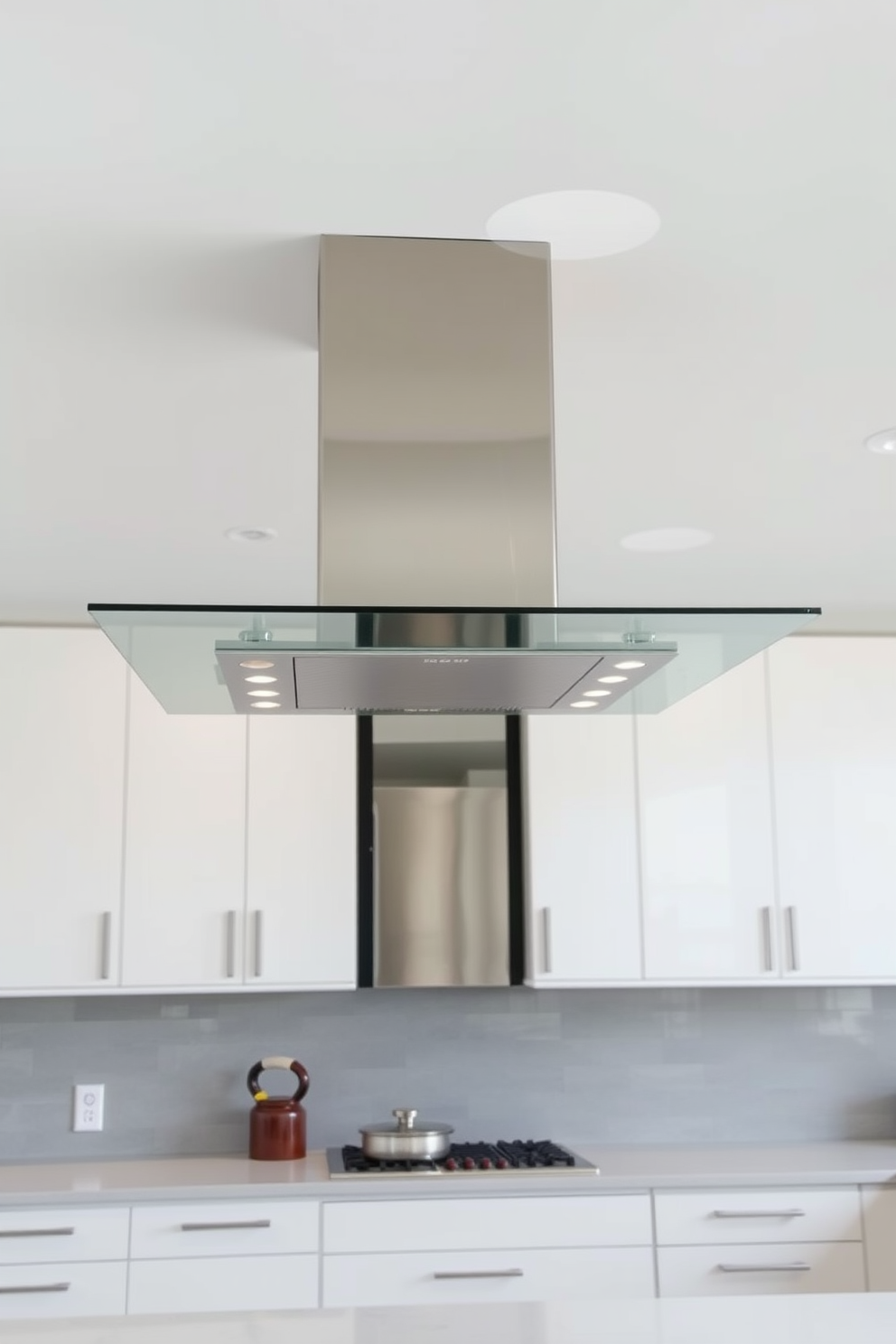 A sleek and modern kitchen hood design featuring a minimalist glass and metal combination. The hood is suspended above a central island, with clean lines and a polished finish that complements the surrounding cabinetry. The glass panel offers a transparent view, allowing light to flow through while providing an elegant touch. The metal accents add a contemporary edge, harmonizing with the overall aesthetic of the kitchen space.