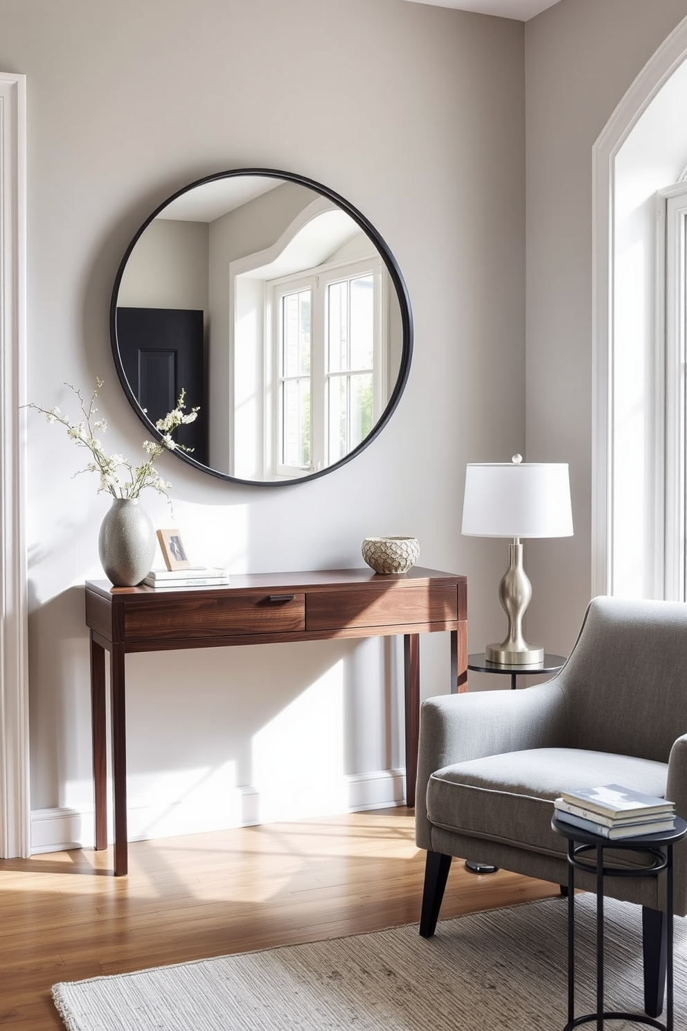 A stylish entryway features a sleek console table made of dark wood with a minimalist design. Above the table, a large round mirror reflects natural light, enhancing the space's openness. To the right of the entryway, a cozy seating area includes a plush armchair upholstered in a soft gray fabric. A small side table beside the chair holds a decorative lamp and a stack of art books, inviting guests to relax.