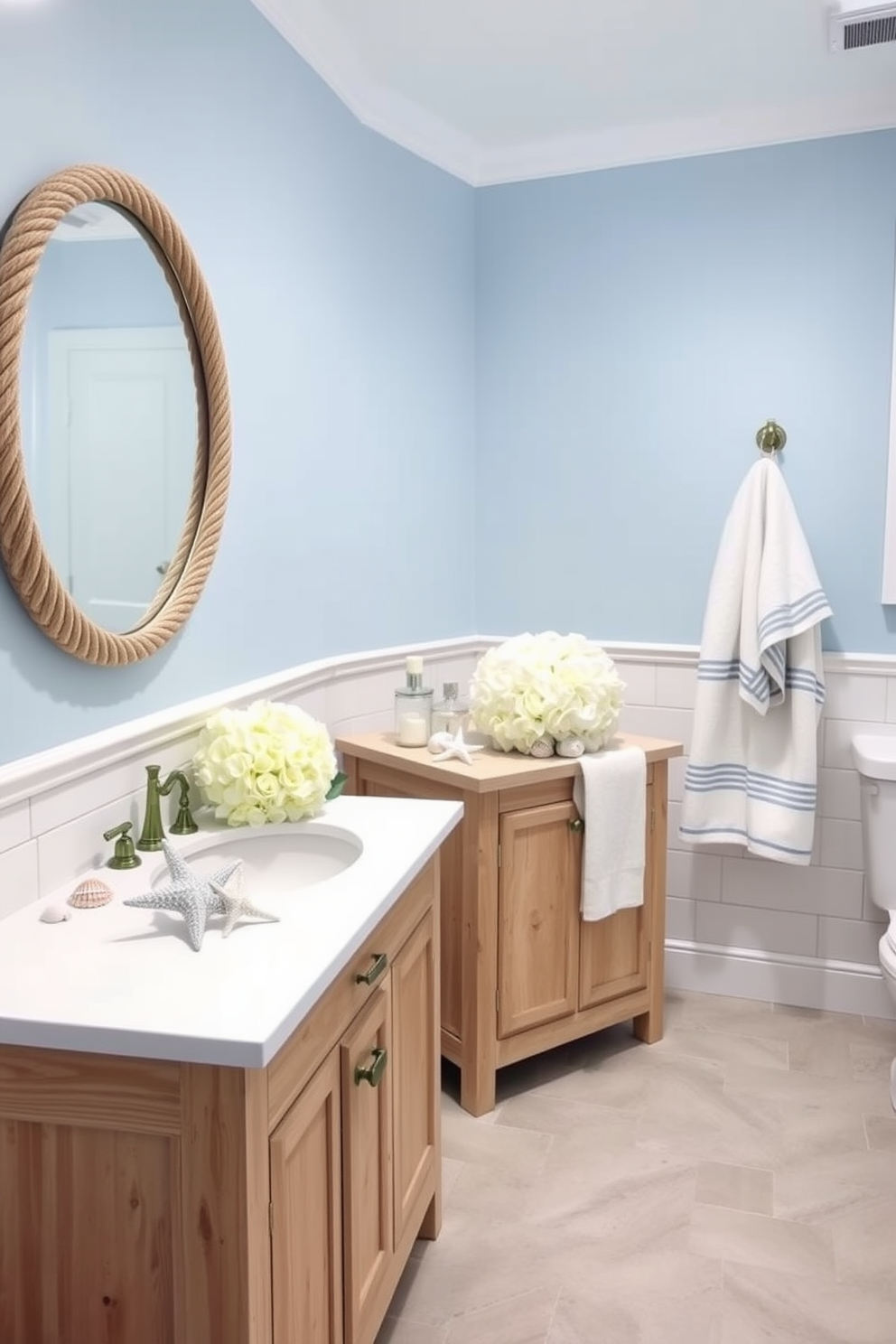 A serene coastal bathroom setting featuring soft blue walls and white shiplap accents. The space includes a wooden vanity with a light driftwood finish and a white quartz countertop, complemented by a large round mirror with a rope frame. Decorative elements include seashells and starfish arranged on the countertop alongside a fresh bouquet of white hydrangeas. The floor is adorned with light gray tiles, and plush towels in nautical stripes hang elegantly from the towel rack.