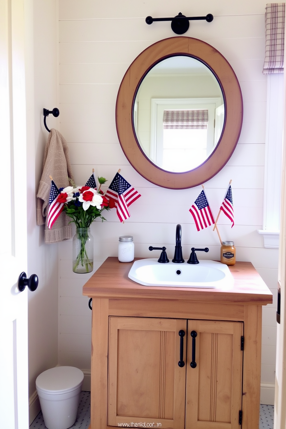 A charming farmhouse style bathroom featuring a rustic wooden vanity with a distressed finish and a white porcelain sink. Above the vanity, a large round mirror with a weathered frame reflects the natural light streaming in from a nearby window. The walls are painted in a soft cream color, complemented by shiplap accents for added texture. Decorate the space with seasonal Labor Day elements like small American flags and a bouquet of red, white, and blue flowers in a mason jar.