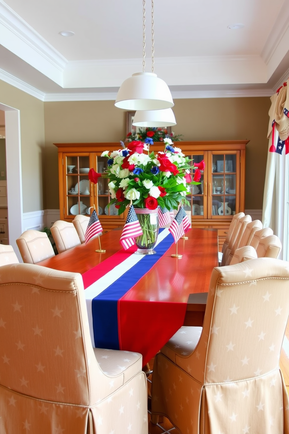 A patriotic dining room setting features a vibrant red white and blue tablecloth draped elegantly over a long wooden dining table. Surrounding the table are matching upholstered chairs adorned with subtle star patterns, creating a festive atmosphere for Labor Day celebrations. The centerpiece is a large glass vase filled with fresh red white and blue flowers, complemented by small American flags placed throughout the table. Soft white pendant lights hang above, casting a warm glow over the scene and enhancing the holiday spirit.