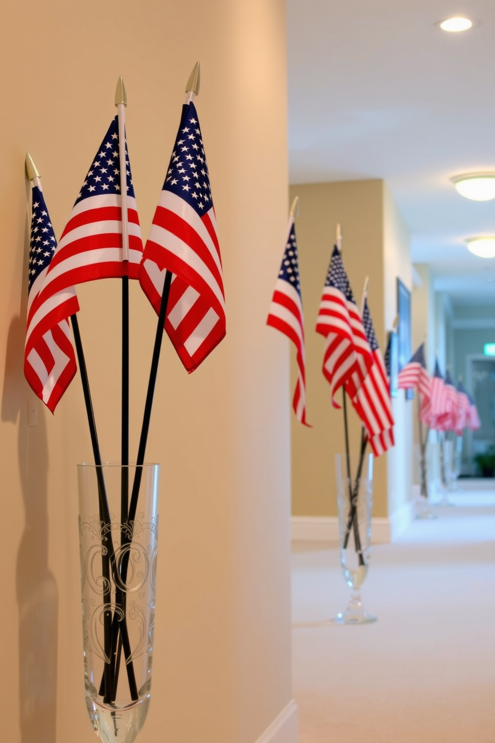 Small American flags are arranged in decorative vases placed along a hallway. The vases are made of clear glass and feature intricate designs, adding elegance to the festive decor. The hallway walls are painted in a soft beige, providing a warm backdrop for the vibrant flags. Subtle lighting fixtures illuminate the space, highlighting the patriotic theme while creating a welcoming atmosphere.