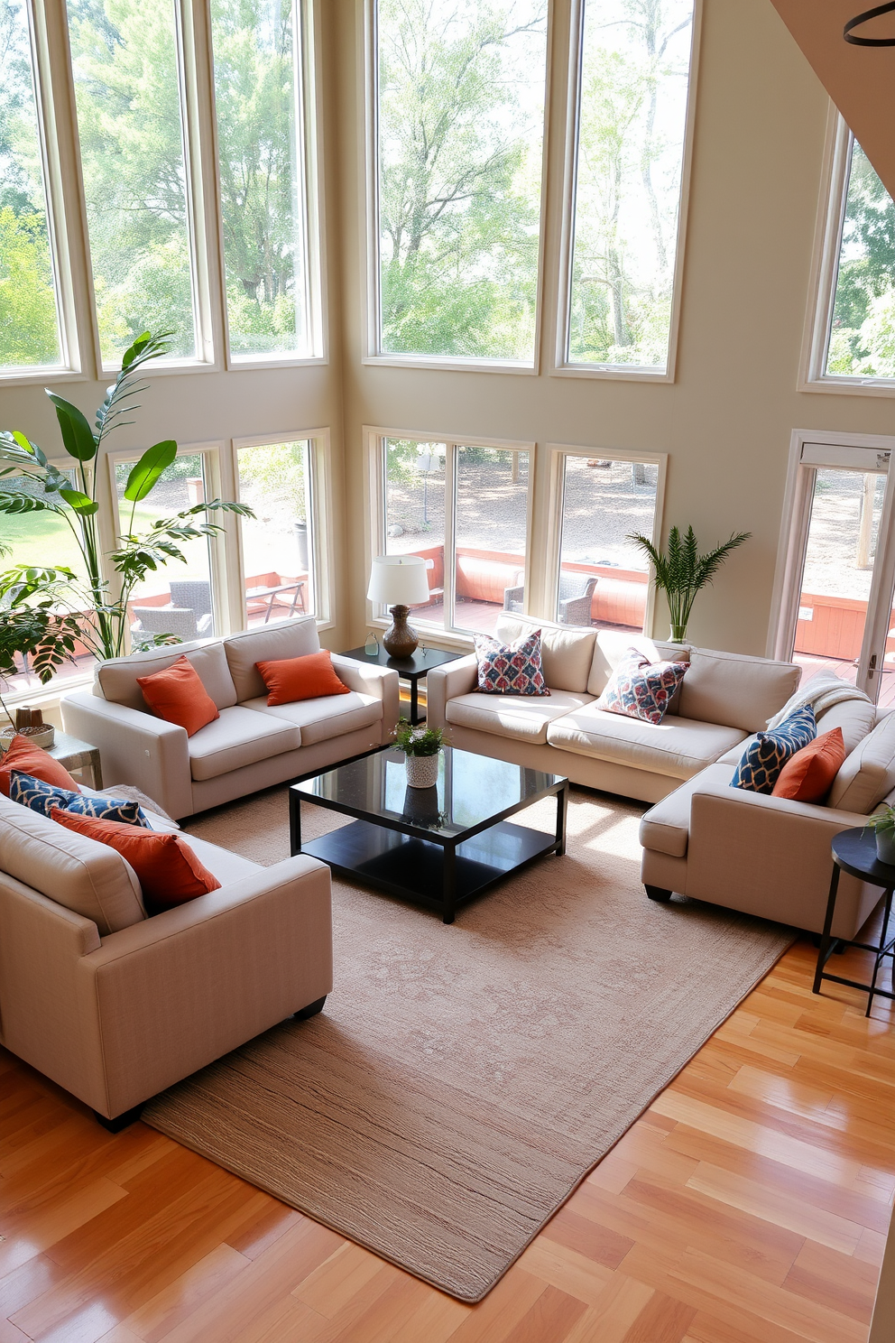 A spacious family room with large windows allowing natural light to flood the space. There are comfortable sectional sofas arranged around a central coffee table, with vibrant throw pillows adding color. In one corner, a tall indoor plant adds a fresh vibe, while a smaller potted plant sits on a side table. The walls are painted in a soft beige, and the hardwood floor is complemented by a large area rug that defines the seating area.