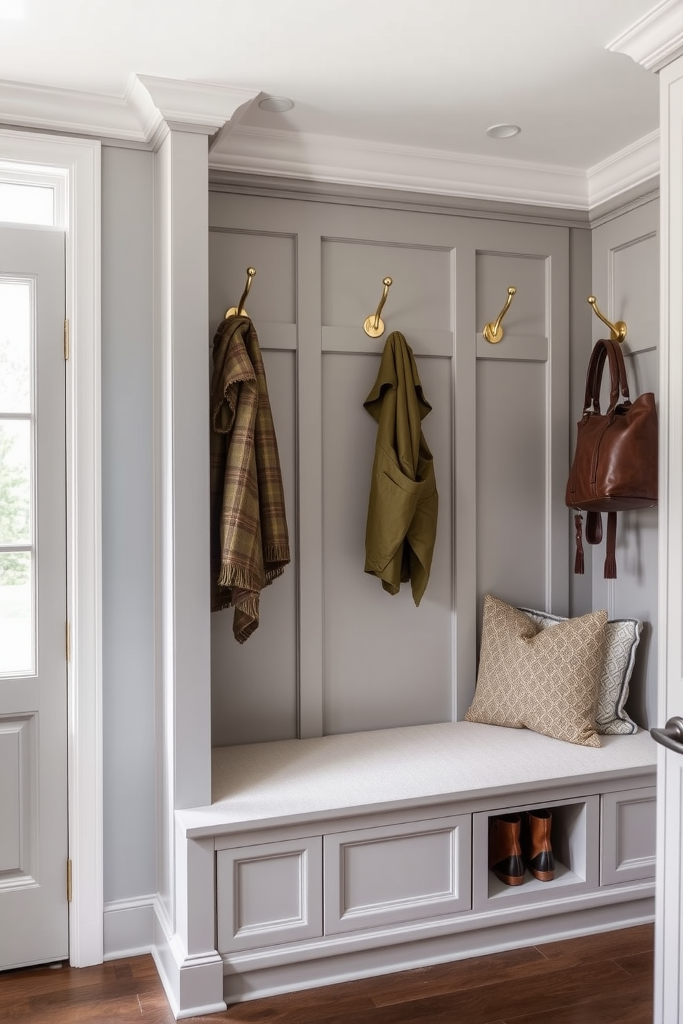 A luxury mudroom features stylish hooks for coats and bags that blend functionality with elegance. The hooks are crafted from polished brass and are mounted on a beautifully paneled wall in a soft gray hue. The space includes a built-in bench with plush cushions and ample storage underneath for shoes and outdoor gear. Natural light floods the room through large windows, enhancing the inviting atmosphere of this sophisticated entryway.