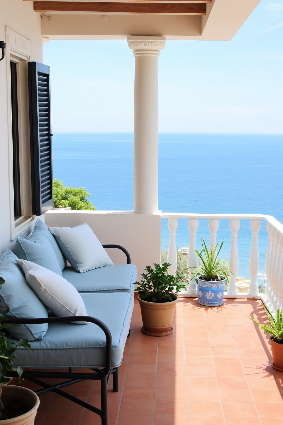 A serene balcony overlooking the ocean features comfortable seating with plush cushions in soft blues and whites. The floor is adorned with terracotta tiles, and potted plants add a touch of greenery, creating a tranquil Mediterranean atmosphere.