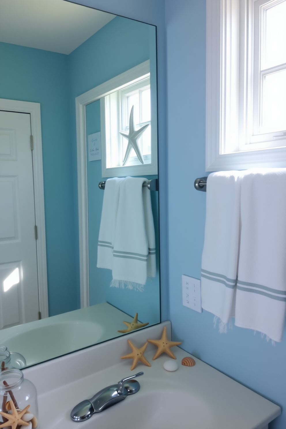 A coastal-themed bathroom featuring decorative starfish and seashell accents. The walls are painted in a soft blue hue, and a large mirror reflects the natural light streaming in from a nearby window. On the countertop, a collection of seashells is arranged in a glass jar, while starfish are scattered artfully around the sink area. White towels with subtle nautical stripes hang neatly on a towel rack, adding to the beach-inspired decor.