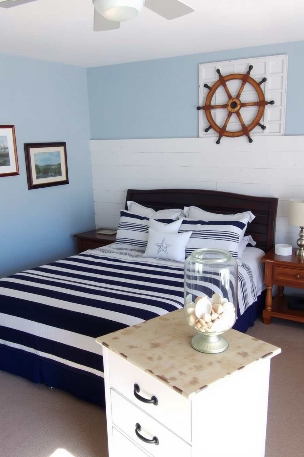 A cozy bedroom featuring nautical themed decor accents. The walls are painted in a soft blue hue, and there are white shiplap panels on one wall adorned with framed maritime artwork. A large bed with a navy and white striped duvet sits in the center, flanked by rustic wooden nightstands. Decorative elements include a vintage ship wheel mounted on the wall and a collection of seashells displayed in a glass jar on the dresser.