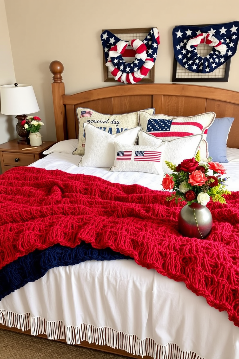 A cozy bedroom setting designed for Memorial Day. The bed is adorned with soft, cozy knitted blankets in red, white, and blue, creating a warm and inviting atmosphere. Decorative pillows with patriotic themes are arranged on the bed. A rustic wooden nightstand holds a small vase filled with fresh seasonal flowers, adding a touch of nature to the decor.