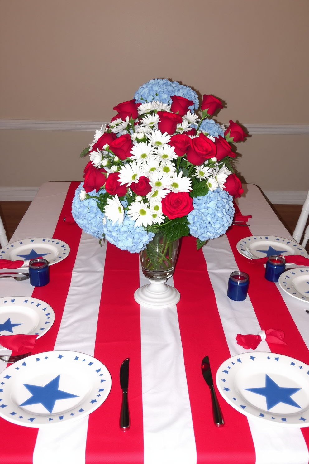 A patriotic themed dining table setting featuring a red, white, and blue tablecloth draped elegantly across the table. Centered on the table is a large floral arrangement with red roses, white daisies, and blue hydrangeas in a classic glass vase. Surrounding the table are white ceramic plates with blue star patterns, complemented by red napkins tied with white ribbon. Candles in red and blue holders are placed evenly along the table, adding a warm glow to the festive atmosphere.