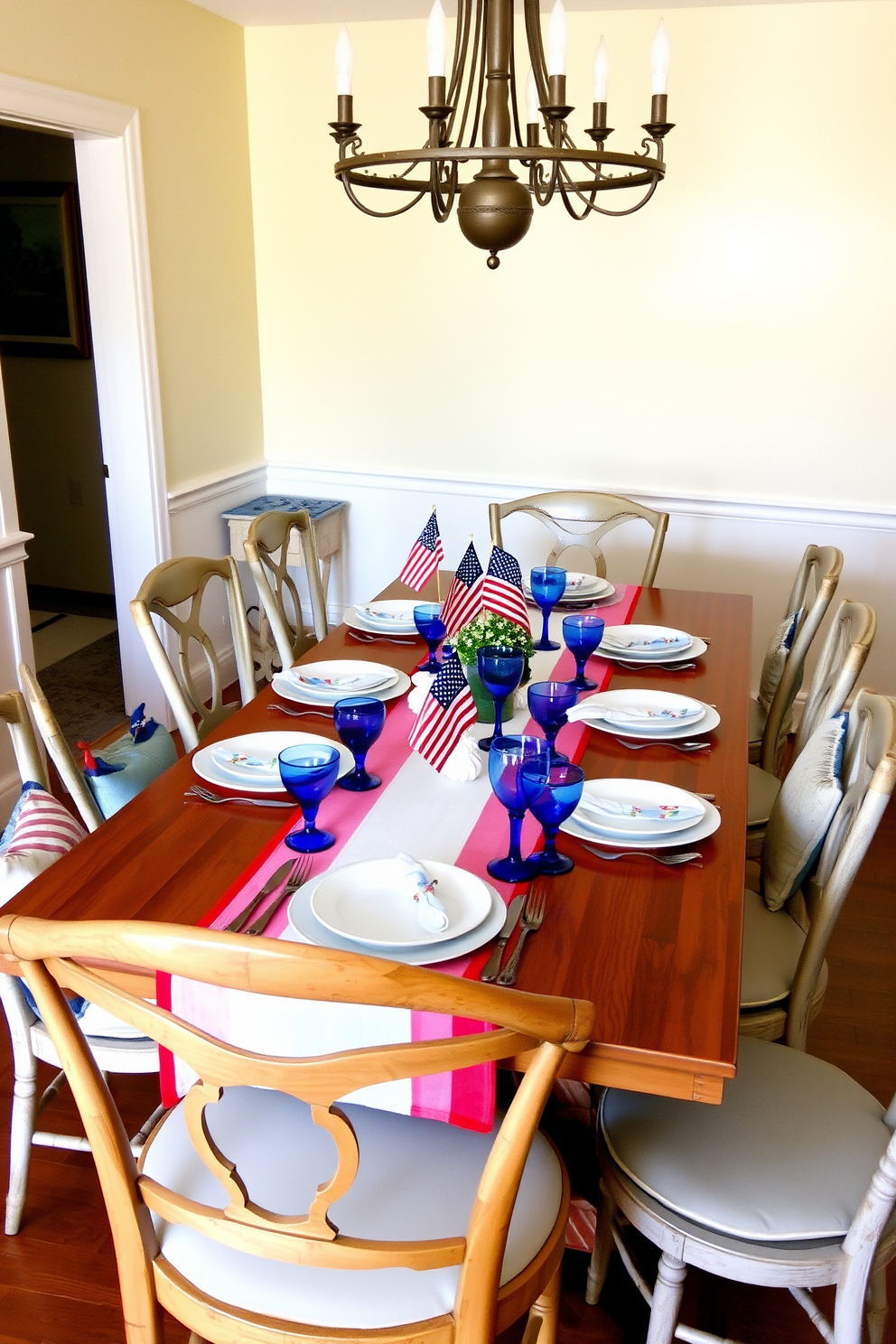 A festive dining room setting featuring a red white and blue table runner that elegantly drapes over a long wooden dining table. Surrounding the table are mismatched vintage chairs, each adorned with soft cushions in complementary colors, creating a warm and inviting atmosphere. On the table, a collection of white dinnerware is arranged alongside vibrant blue glassware, while small American flags serve as charming centerpieces. The walls are painted in a soft cream color, and a rustic chandelier hangs overhead, casting a warm glow over the festive decor.