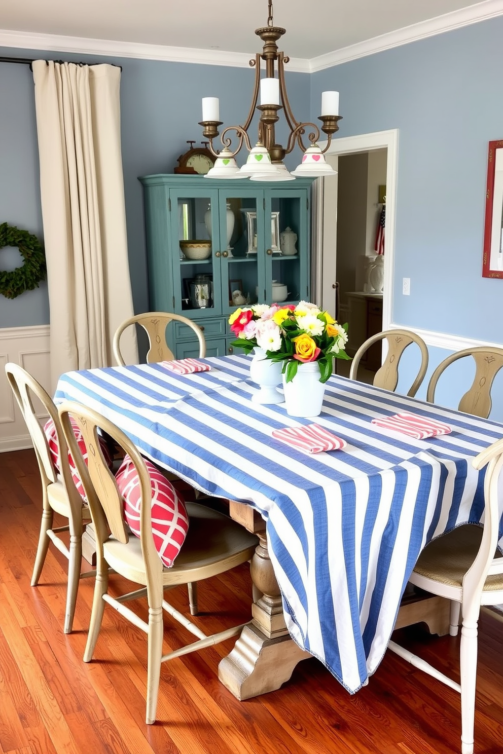 A charming dining room featuring a blue and white striped tablecloth draped elegantly over a rustic wooden table. Surrounding the table are mismatched vintage chairs, each adorned with a small decorative pillow in coordinating colors. On the table, a centerpiece of fresh flowers in a white vase adds a touch of brightness, complemented by red and white striped napkins. Soft lighting from a hanging chandelier casts a warm glow over the space, enhancing the festive atmosphere for Memorial Day.