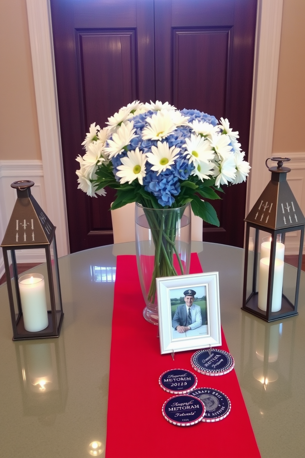 An elegant entry table adorned with themed centerpieces celebrating Memorial Day. The table is decorated with a vibrant red white and blue runner and features a large glass vase filled with fresh white daisies and blue hydrangeas. Flanking the vase are two decorative lanterns with flickering candles inside creating a warm inviting glow. A small framed photo of a military veteran stands prominently on the table alongside a few patriotic themed coasters.