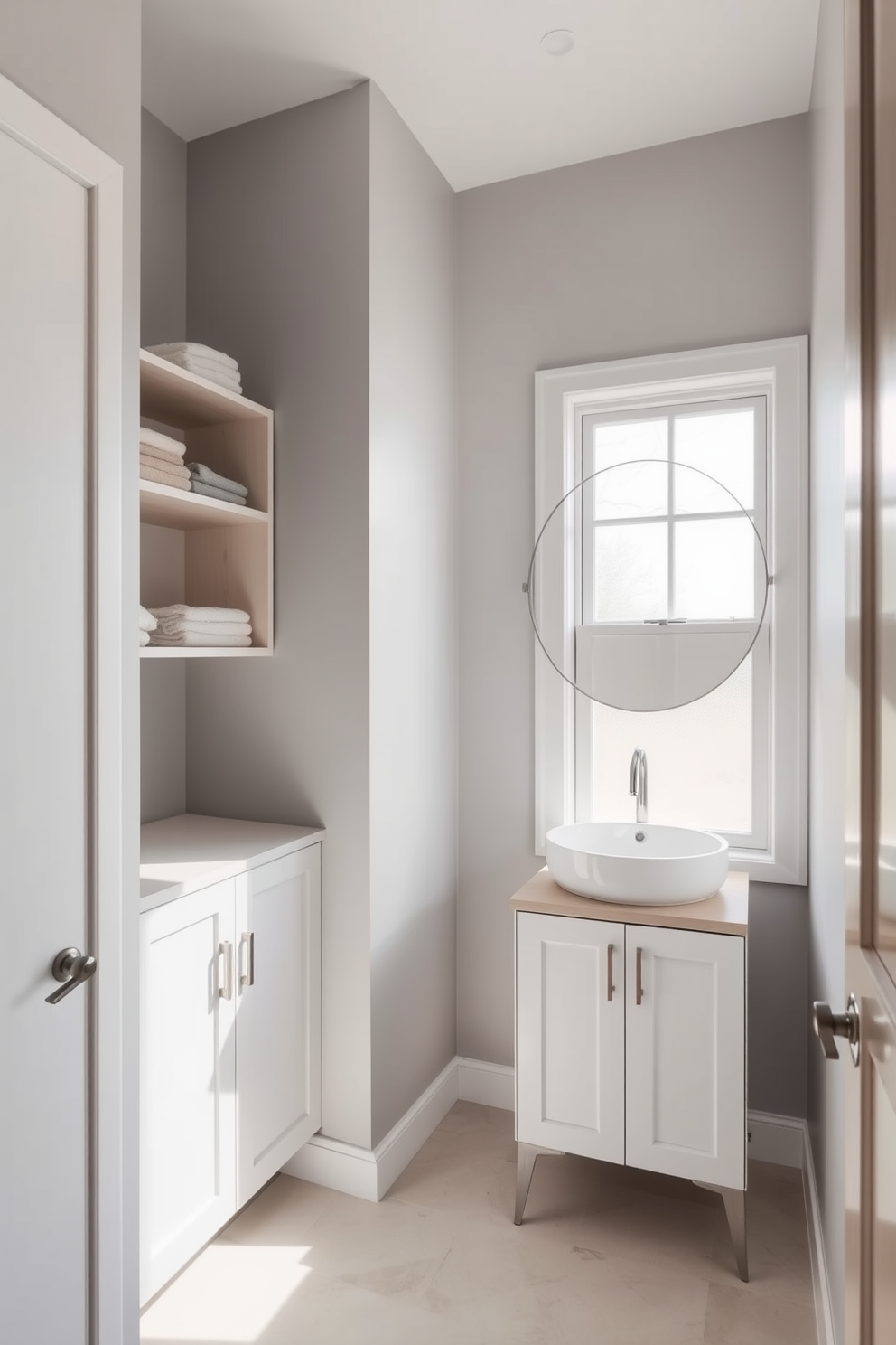 A minimalist bathroom featuring compact storage solutions designed to reduce clutter. The space includes sleek floating shelves and a built-in cabinet, both painted in a soft white finish to enhance the airy feel. Natural light floods the room through a large frosted window, illuminating the simple yet elegant fixtures. The walls are painted in a calming pale gray, complemented by a light wood vanity with a single sink and a large round mirror above it.