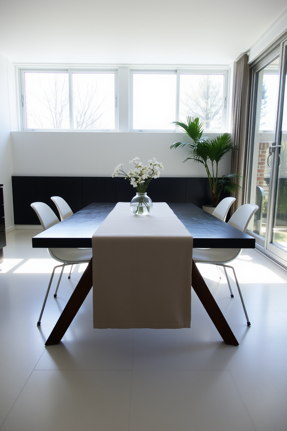 A minimalist dining room features a simple runner on the dining table that complements the overall aesthetic. The table is surrounded by sleek, modern chairs, and natural light streams in through large windows, creating an inviting atmosphere.