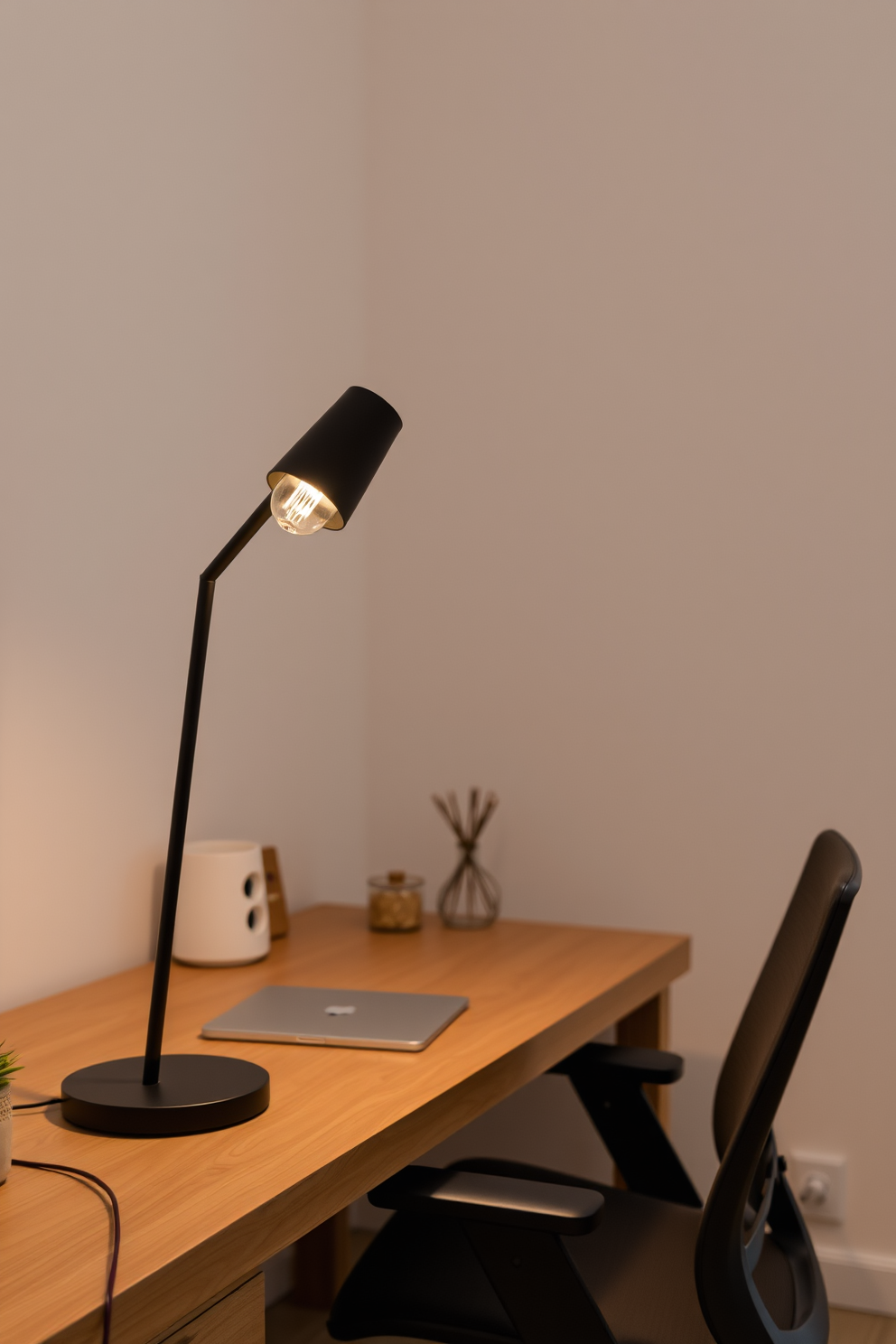 A stylish desk lamp with a sleek design illuminates a minimalist workspace. The lamp features a matte black finish and a warm white bulb, creating a cozy yet modern atmosphere. The home office is adorned with a simple wooden desk and a comfortable ergonomic chair. Neutral tones dominate the color palette, complemented by a few carefully chosen decorative elements.