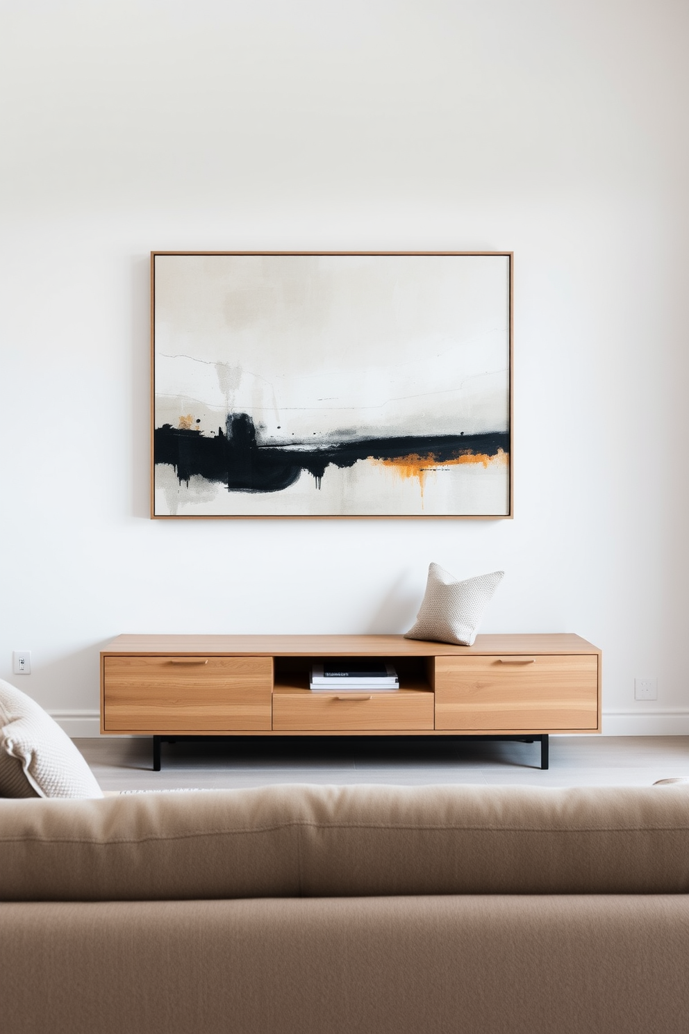 A minimalist living room featuring a low-profile media console in a sleek, modern style. The console is finished in a light oak wood and sits against a soft white wall, complementing the overall serene aesthetic of the space. A plush, neutral-toned sofa faces the console, adorned with a few textured throw pillows. A large abstract painting hangs above the sofa, adding a pop of color and visual interest to the room.