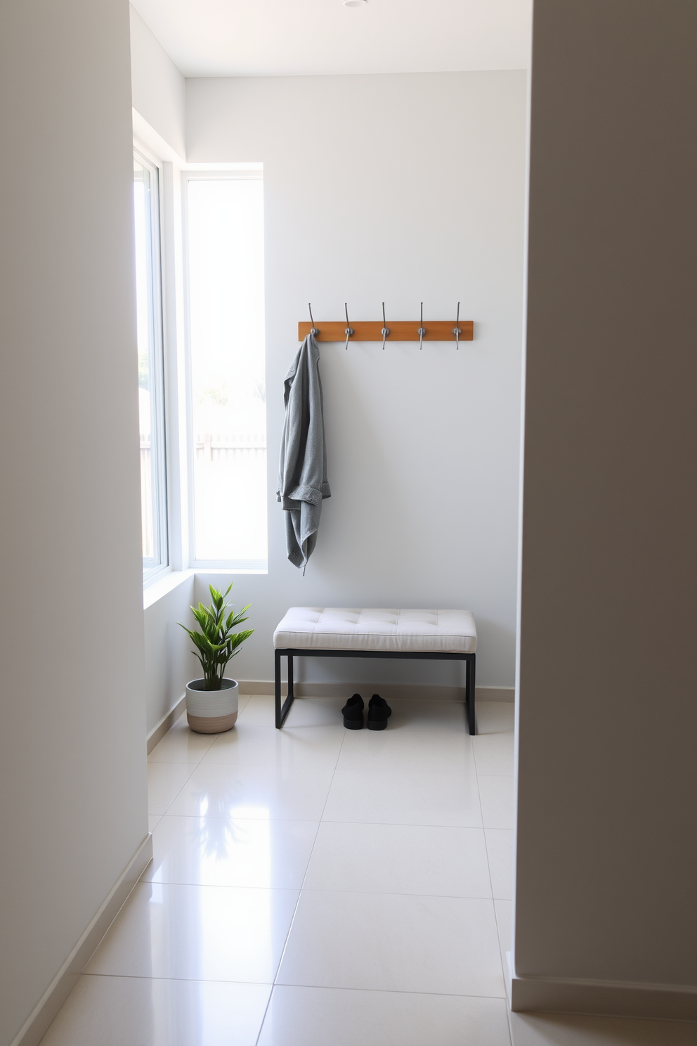 A minimalist coat rack with sleek wooden hooks stands against a light gray wall. Below the rack, a simple bench with a soft cushion invites guests to sit and remove their shoes. The floor is adorned with large light-colored tiles that reflect natural light streaming in from a nearby window. A small potted plant sits in the corner, adding a touch of greenery to the modern entryway.