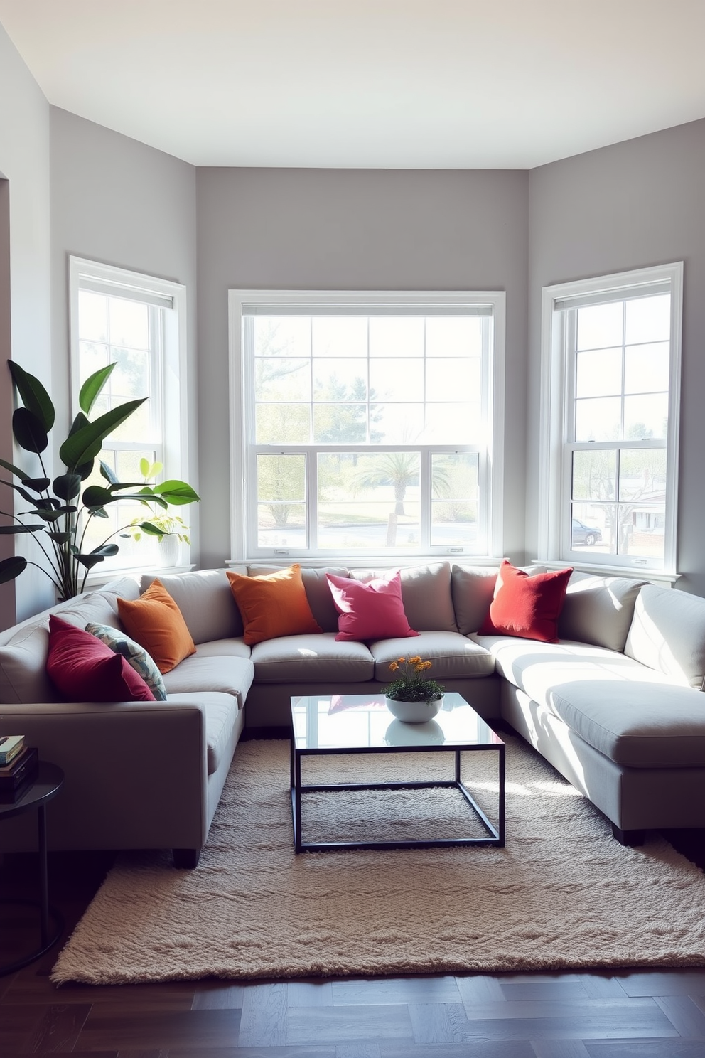 A modern family room filled with natural light. The space features a large sectional sofa in a neutral tone, complemented by vibrant throw pillows and a sleek coffee table. In one corner, a tall indoor plant adds a touch of greenery, while a smaller potted plant sits on a side table. The walls are painted in a soft gray, and a cozy area rug anchors the seating arrangement.