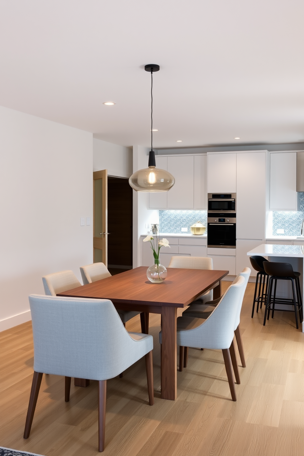 A minimalist dining area features a simple wooden table surrounded by sleek, upholstered chairs. The space is illuminated by a pendant light that adds a warm ambiance to the neutral-colored walls. In the modern kitchen design, clean lines and a functional layout are emphasized with high-end appliances and a large island. The cabinetry is finished in a matte white, complemented by a striking backsplash that adds a touch of personality.