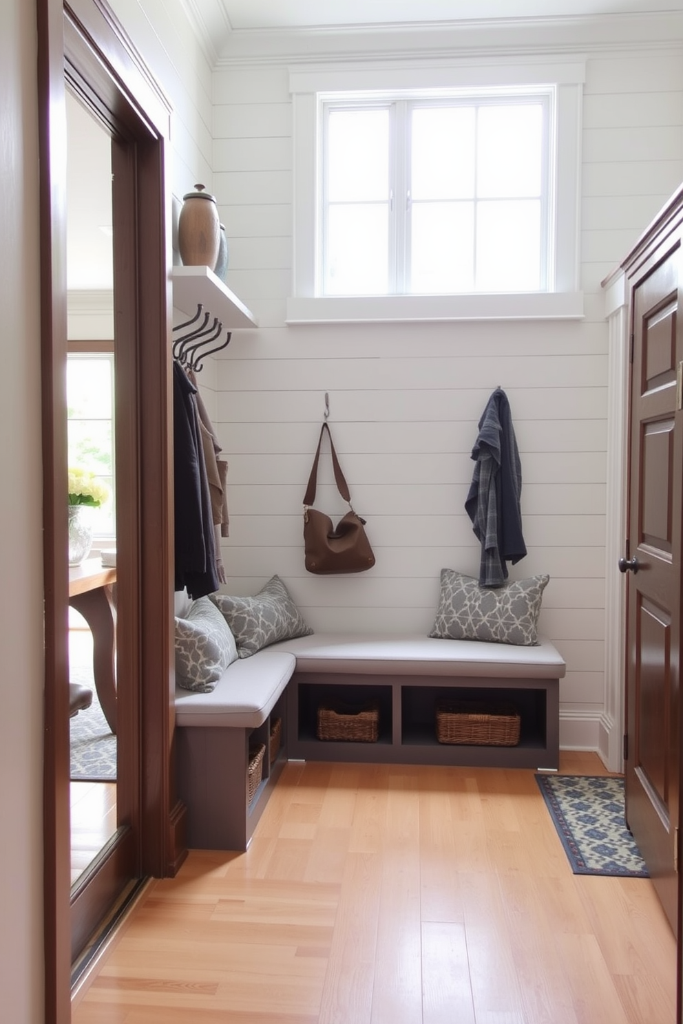 A stylish mudroom featuring a built-in bench with ample storage underneath. The bench is upholstered in a soft gray fabric, and above it, there are hooks for coats and bags, creating an organized and welcoming space. Adjacent to the dining room, the mudroom flows seamlessly with a cohesive color palette. Light wood flooring complements the white shiplap walls, while a large window allows natural light to brighten the area.