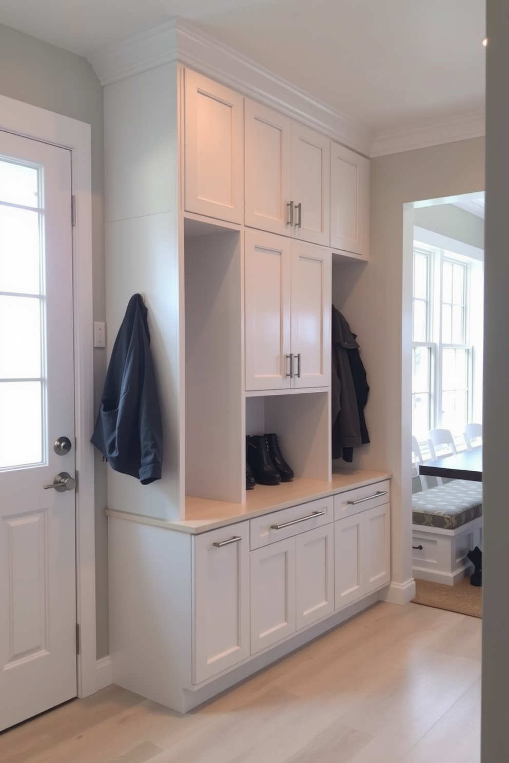 A stylish mudroom featuring built-in cabinets for a sleek look. The cabinets are finished in a soft white with brushed nickel hardware, providing ample storage for shoes and outerwear. Adjacent to the dining room, the mudroom is designed with a functional bench for seating. Natural light floods the space through a nearby window, enhancing the inviting atmosphere.