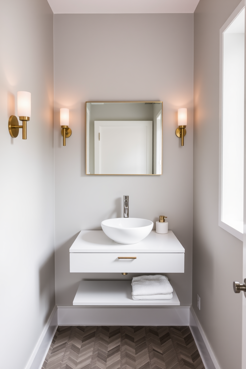 A narrow powder room features a sleek floating vanity with a white countertop and a stylish vessel sink. The walls are painted in a soft gray hue, complemented by elegant wall sconces that provide warm lighting. A large rectangular mirror hangs above the vanity, reflecting the chic decor. The floor is adorned with herringbone-patterned tiles in muted tones, adding depth to the compact space.