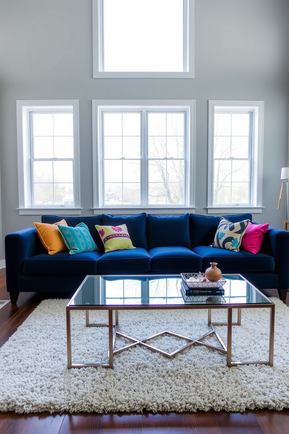 Light gray walls create a serene backdrop for a living room featuring a navy blue couch. The couch is adorned with an array of colorful throw pillows, providing a striking contrast against the muted wall color. A sleek coffee table made of glass and metal sits in front of the couch, complemented by a plush area rug underneath. Floor-to-ceiling windows allow natural light to flood the space, enhancing the inviting atmosphere.