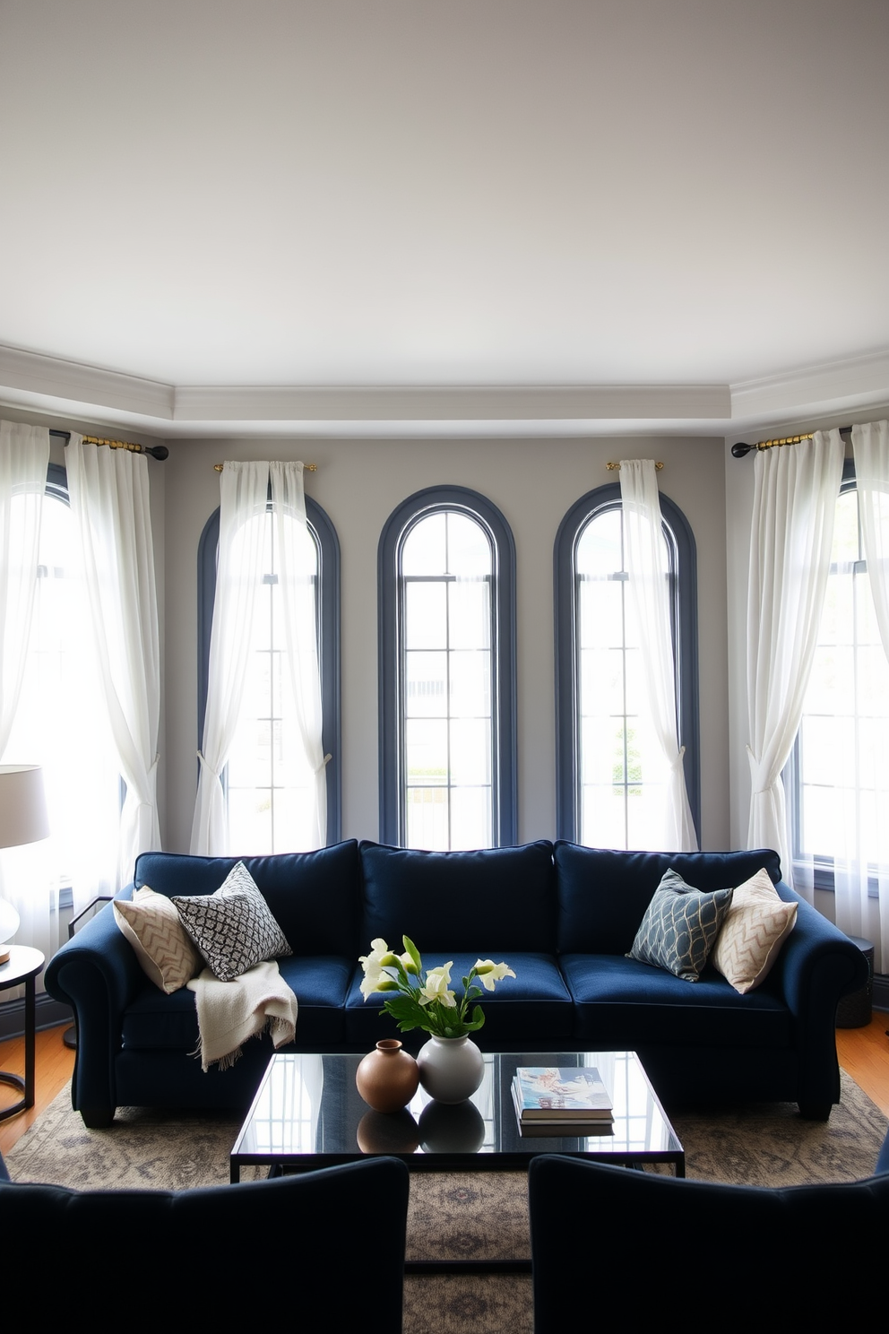 A cozy living room featuring a navy blue couch that serves as the focal point of the space. The room is adorned with white curtains that elegantly frame the large windows, allowing natural light to flood in. The walls are painted in a soft gray tone, creating a serene backdrop for the vibrant couch. A stylish coffee table sits in front of the couch, complemented by decorative pillows in various textures and patterns.