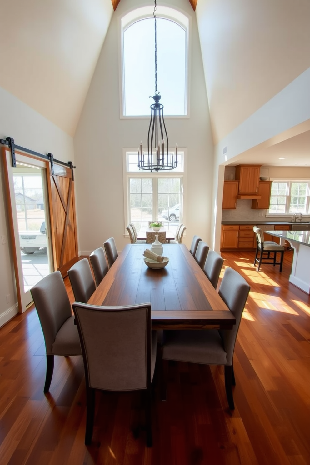 A spacious open floor plan dining room features a large wooden dining table surrounded by upholstered chairs. Natural light floods the space through oversized windows, enhancing the warm tones of the hardwood flooring. In one corner, a sliding barn door adds rustic charm and separates the dining area from the adjacent kitchen. The walls are painted in a soft neutral color, while a statement chandelier hangs above the table, creating an inviting atmosphere.