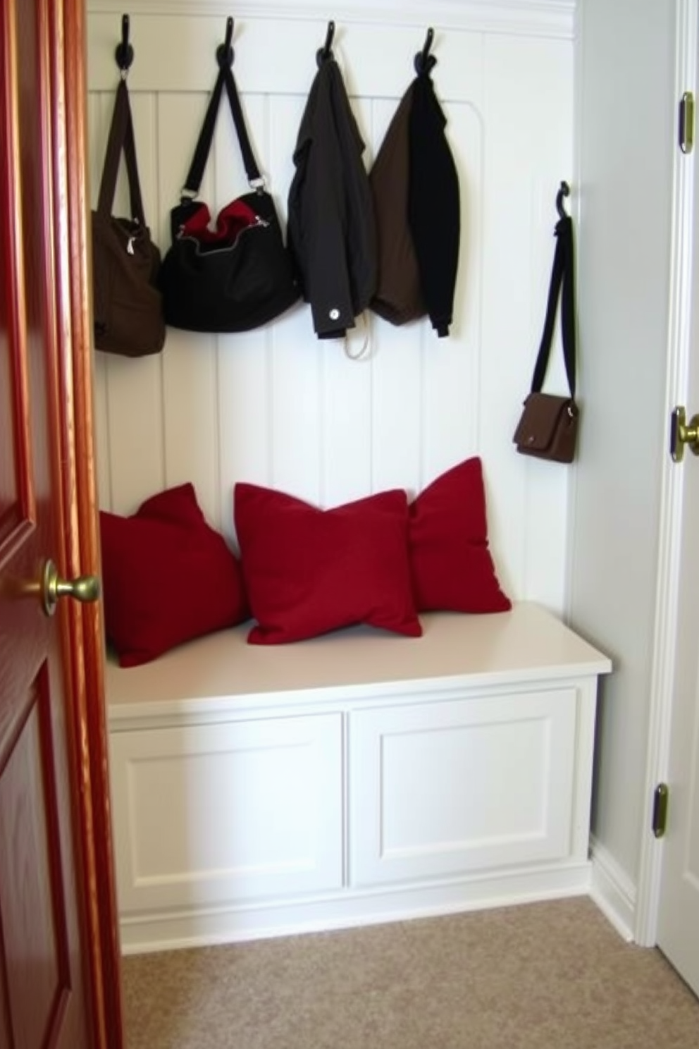 A cozy mudroom designed with functionality in mind. The space features a built-in bench with storage underneath, surrounded by hooks for coats and bags. Red decorative pillows are strategically placed on the bench for added comfort and warmth. The walls are painted in a soft white, creating a bright and inviting atmosphere.