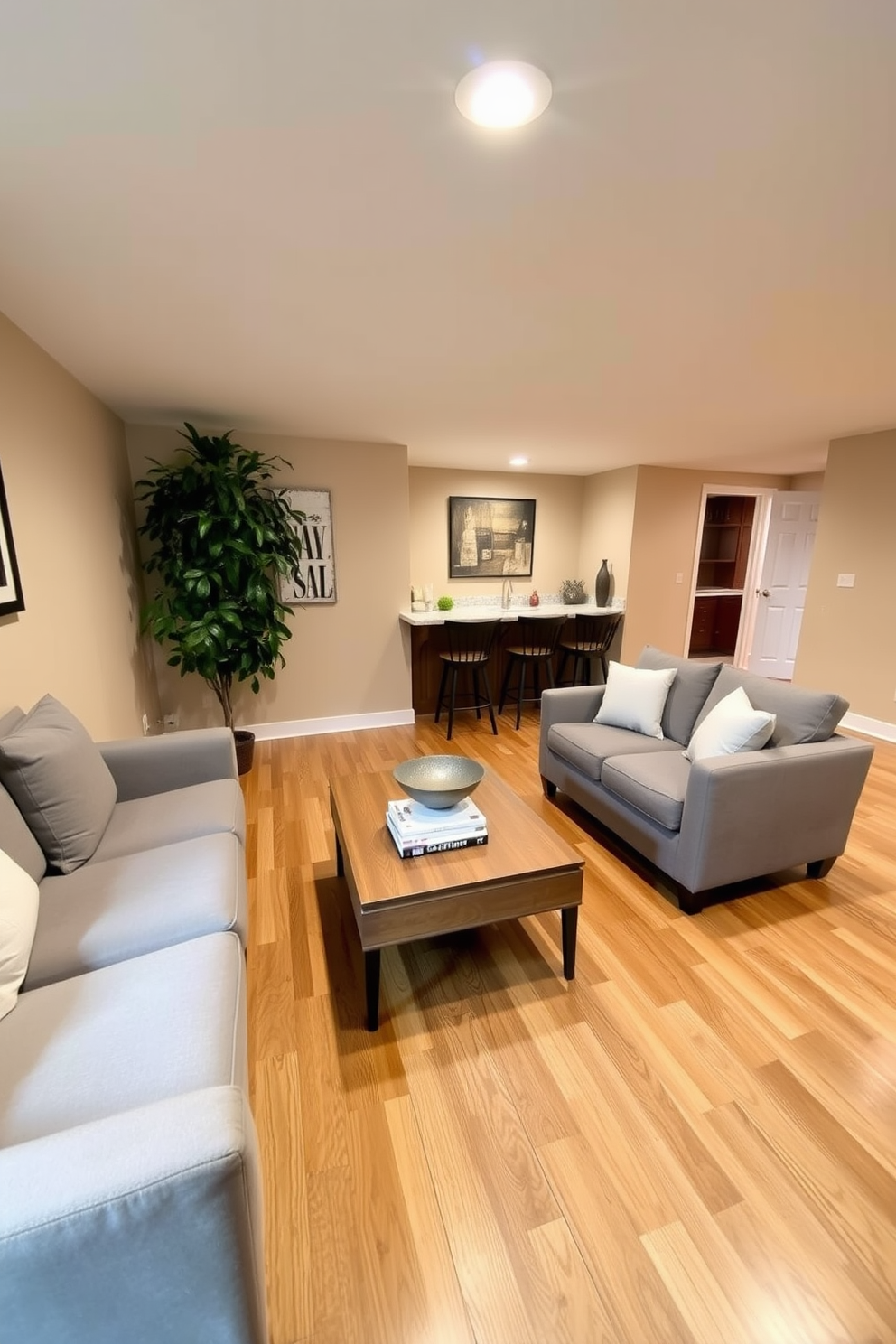 A cozy basement space featuring a comfortable sectional sofa in soft gray fabric. The walls are painted in a light beige color, enhancing the brightness of the area. A wooden coffee table sits in the center, adorned with a stack of books and a decorative bowl. In one corner, a tall indoor plant adds a refreshing touch of greenery to the room. The flooring is a warm oak laminate, providing a welcoming feel. A small bar area with a few stools is positioned against one wall, perfect for casual gatherings.