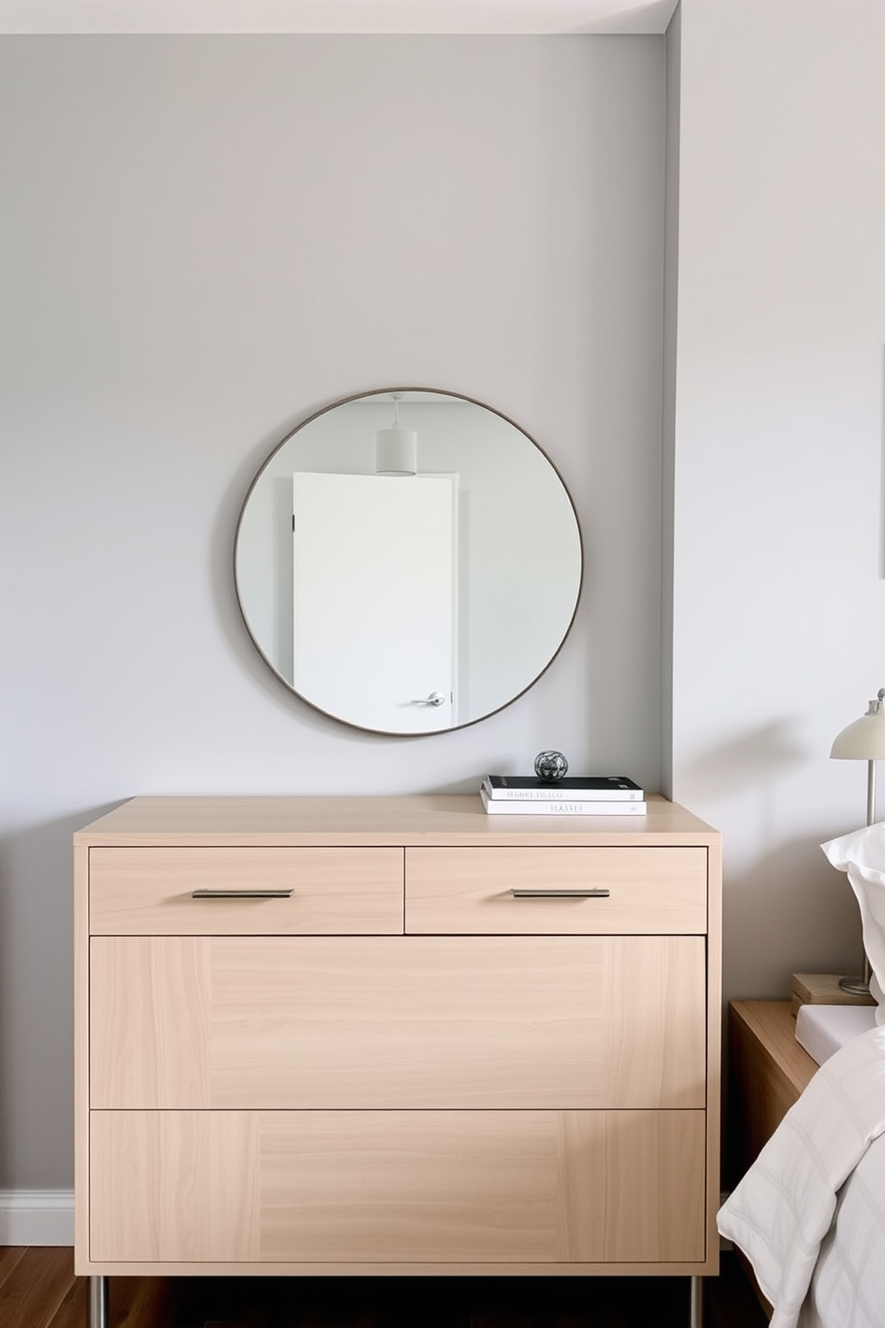 A functional dresser with a stylish design stands against a soft gray wall in a cozy bedroom. The dresser features clean lines and a light wood finish, complemented by sleek metal handles and a large round mirror above it. The bed, dressed in crisp white linens, is positioned next to the dresser, creating a serene atmosphere. A small bedside table with a minimalist lamp adds to the simplicity of the overall bedroom design.