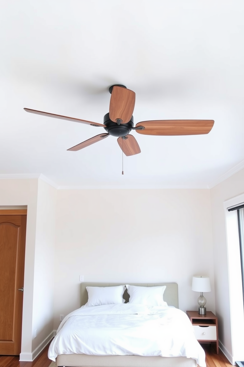 A simple ceiling fan with a sleek design is mounted in the center of the room, providing gentle air circulation. The fan features wooden blades that complement the soft color palette of the space. The bedroom is designed with a minimalist approach, featuring a low-profile bed with crisp white linens. A small nightstand with a modern lamp sits beside the bed, while a cozy area rug adds warmth to the hardwood floor.