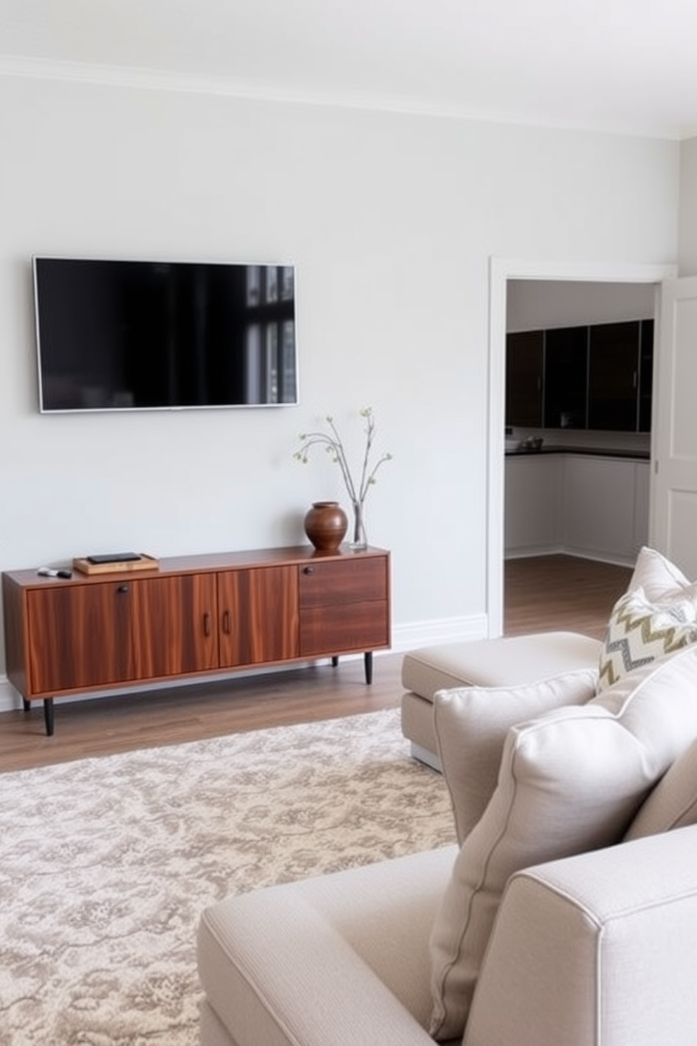 A contemporary family room featuring a wall-mounted television above a sleek media console. The console is finished in a rich walnut wood, providing a warm contrast to the light gray walls. Cozy seating arrangements include a plush sectional sofa adorned with decorative throw pillows. A soft area rug anchors the space, complementing the overall minimalist aesthetic.