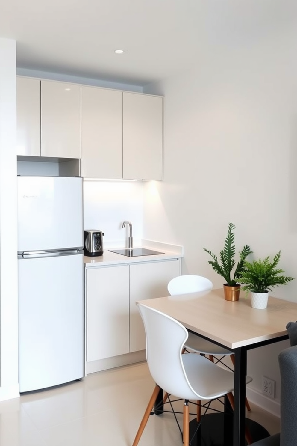 A slim refrigerator is positioned against the wall, maximizing the available space in the small apartment kitchen. The kitchen features sleek cabinetry in a light color, complemented by a minimalist countertop that enhances the open feel of the area. A compact dining table with modern chairs is placed nearby, creating a cozy dining nook without overcrowding the space. The walls are painted in a soft white, and a few decorative plants add a touch of greenery, making the kitchen inviting and functional.