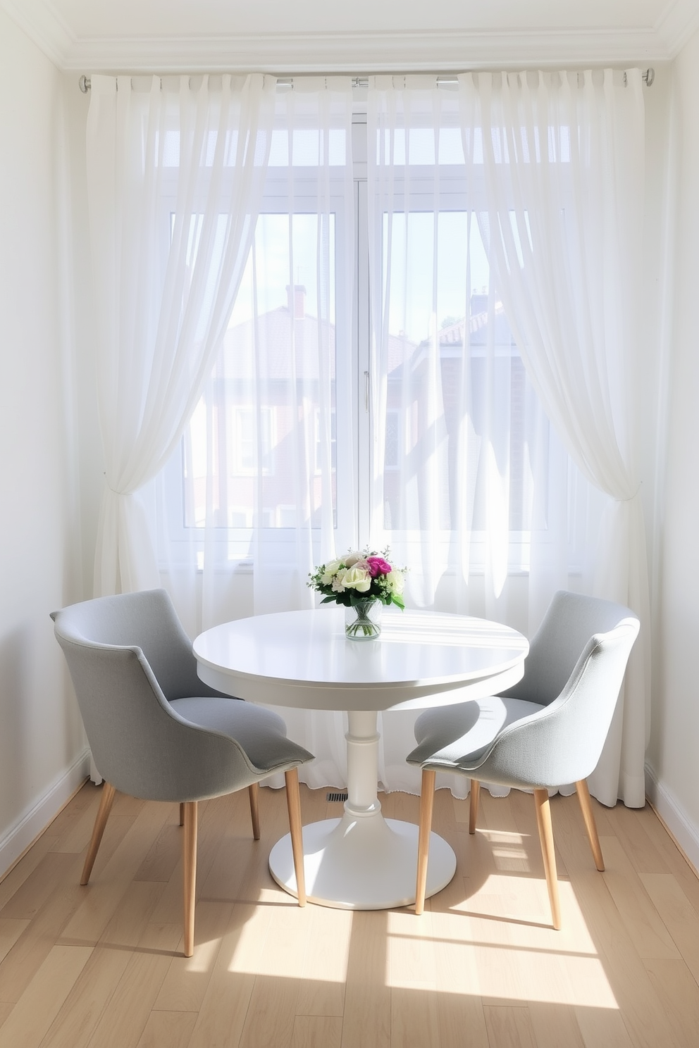 A small dining room features a round white table surrounded by light gray upholstered chairs. Soft natural light filters in through sheer white curtains, illuminating the space and creating an airy atmosphere. The walls are painted in a soft pastel hue, complementing the light wood flooring. A simple centerpiece of fresh flowers adds a touch of elegance to the table while maintaining a minimalist aesthetic.